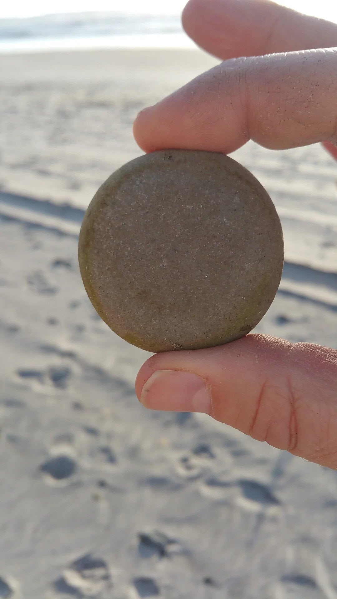 Hand holding a round, smooth stone on the beach with sand and ocean in the background.