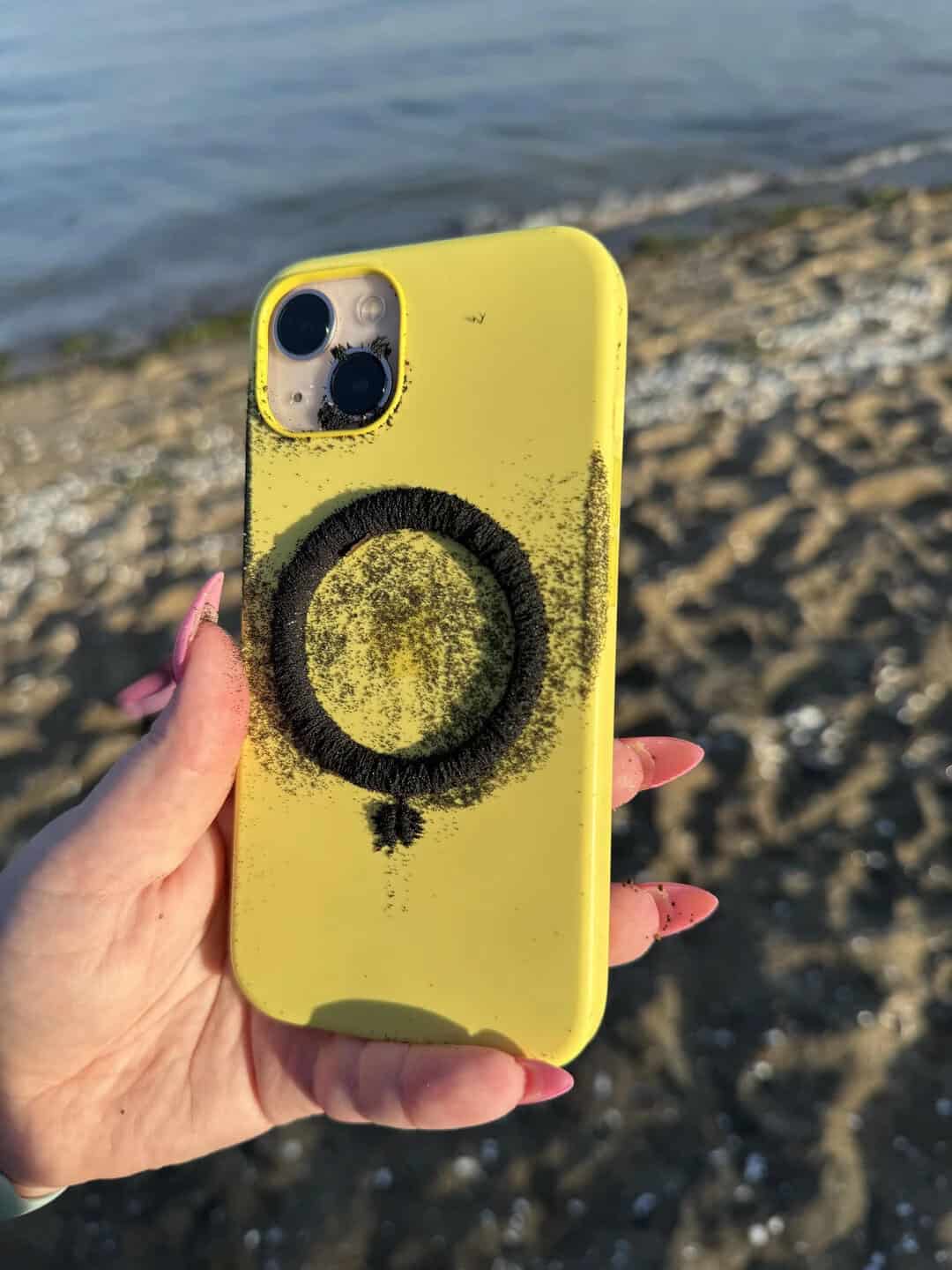 Yellow phone with black ring design held by hand on beach with water in background.