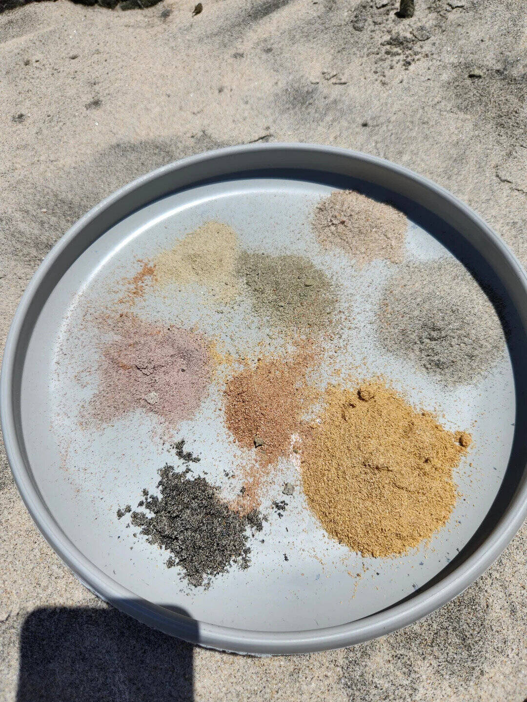 Collection of various colored sands on a round tray at the beach.