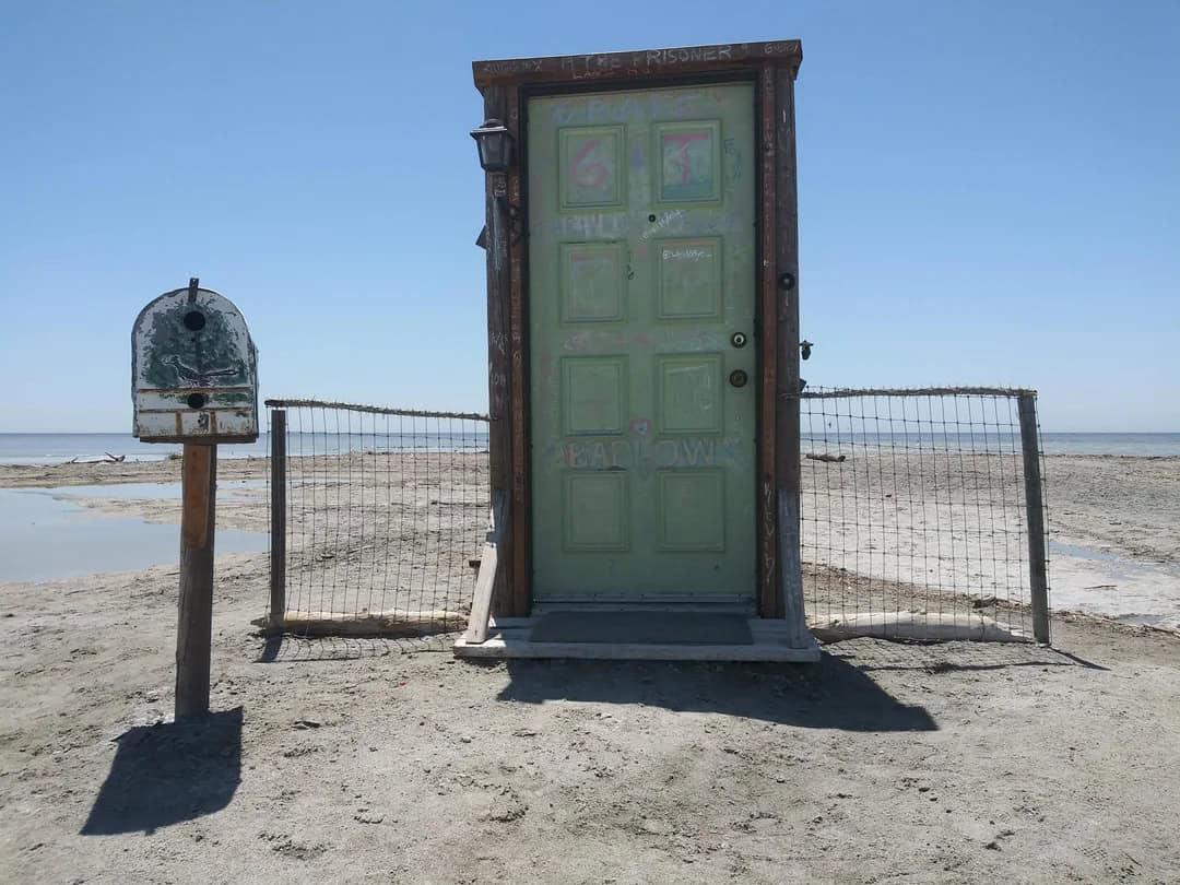 A green door with graffiti on a beach, surrounded by a fence and a mailbox.