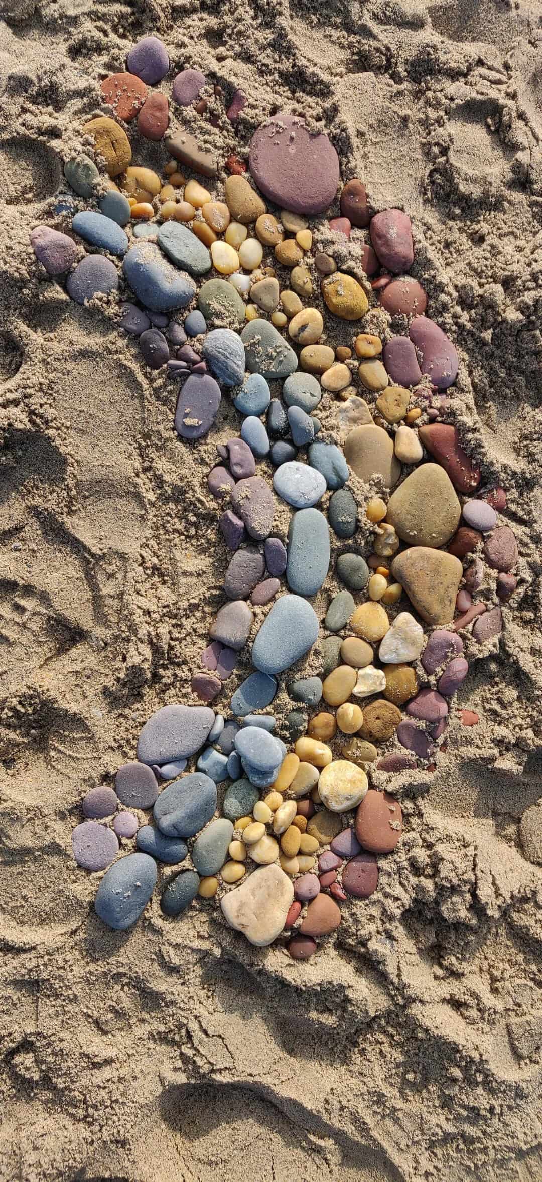 Colorful pebbles arranged on sandy beach with footprints nearby.