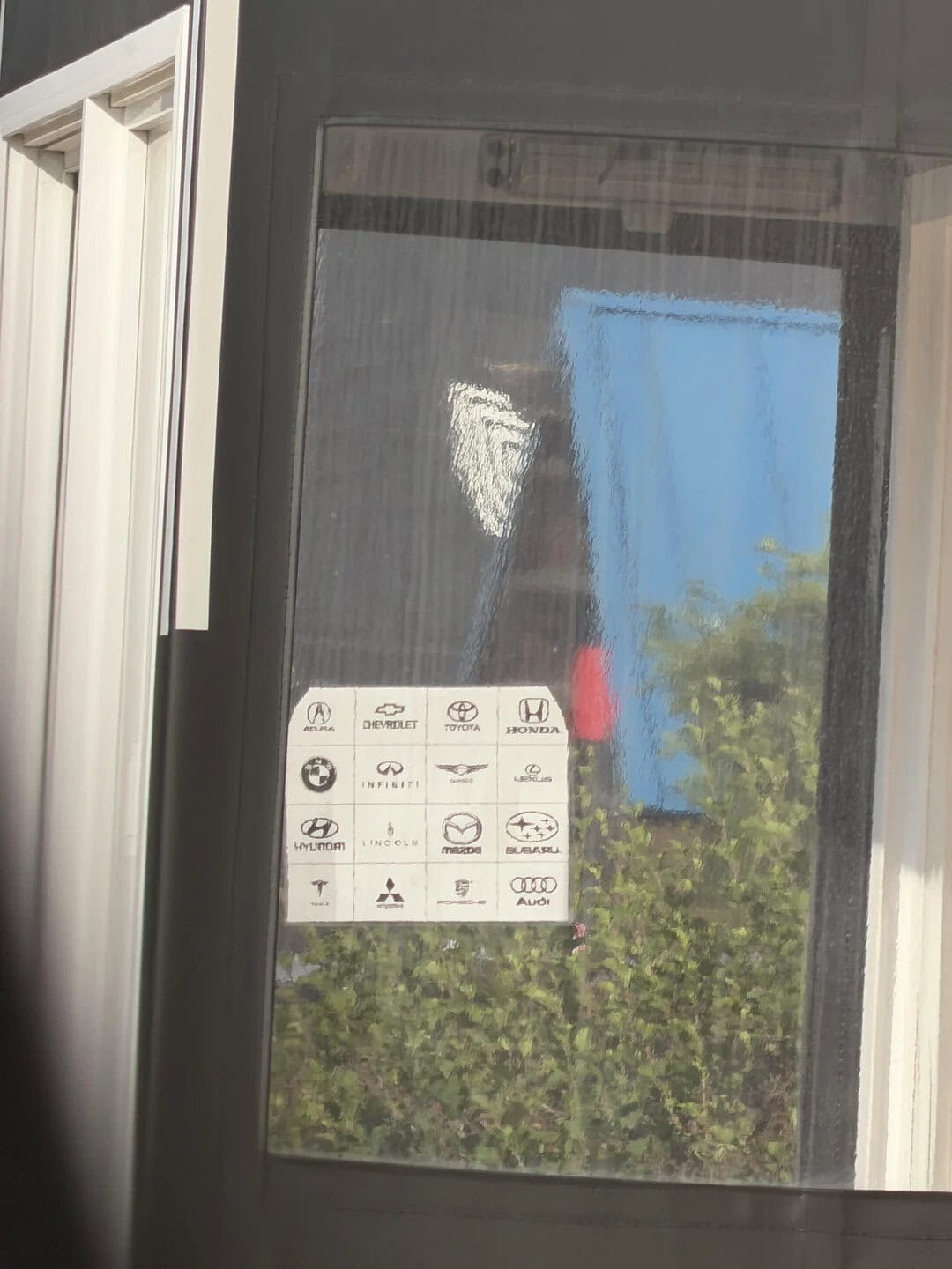Reflection of a blue sky and trees in a glass window with a white sign displaying various car brand.