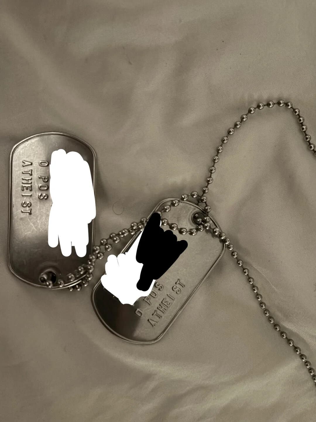 Two silver dog tags with blacked-out text on a ball chain necklace on a beige fabric background.