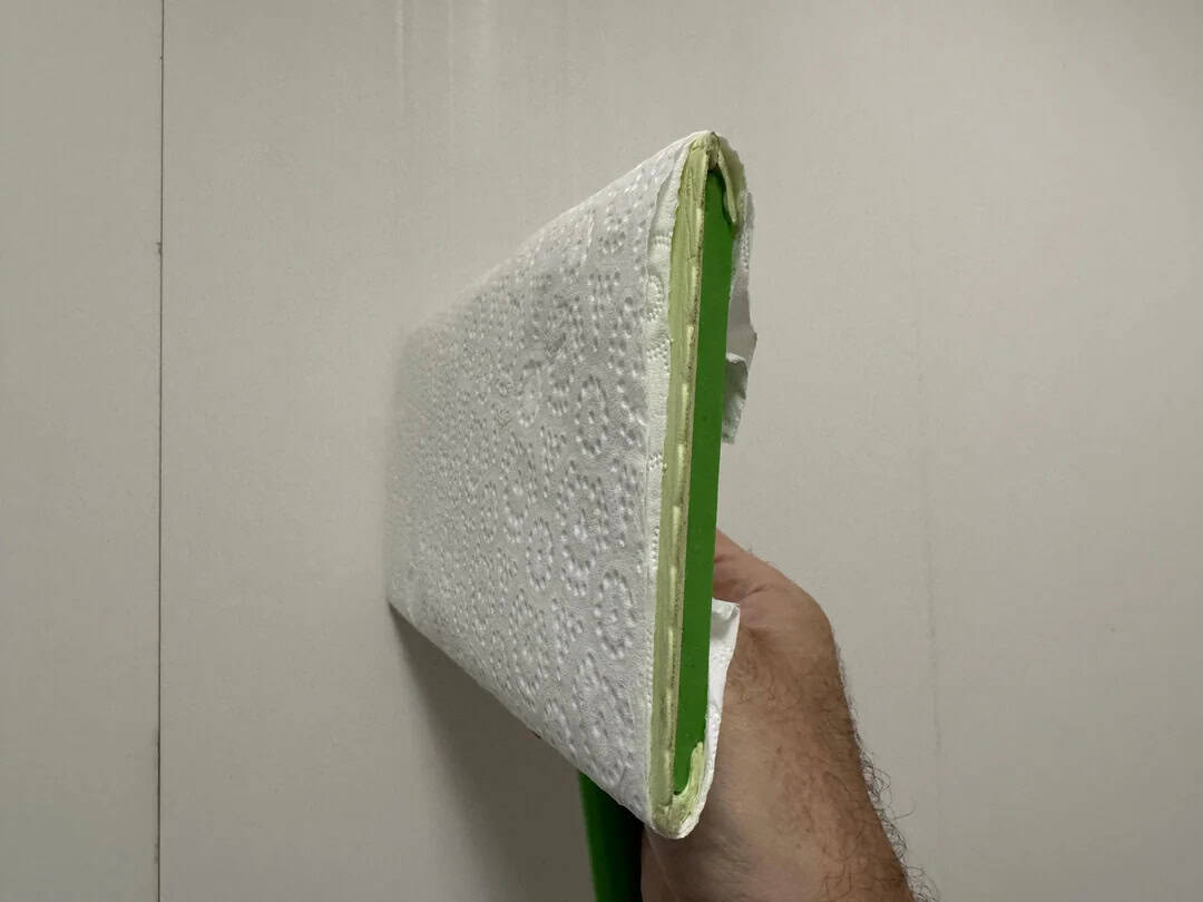 Hand holding a green and white disposable mop head against a plain wall.