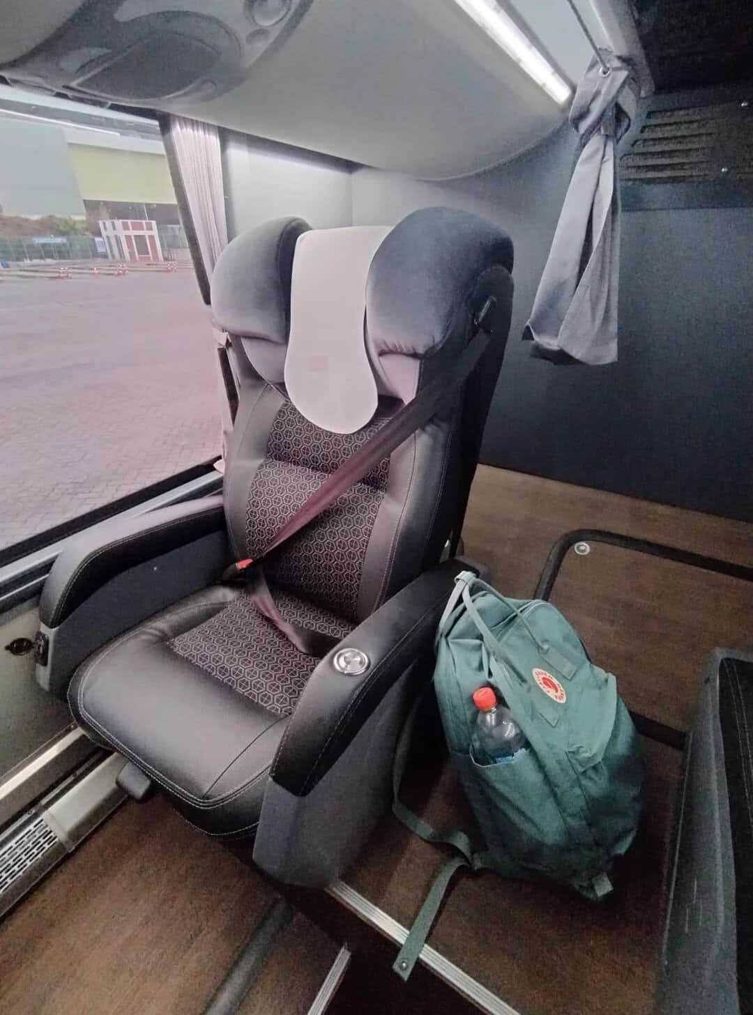Child car seat with headrest and harness, next to a backpack with a water bottle inside, on a bus.