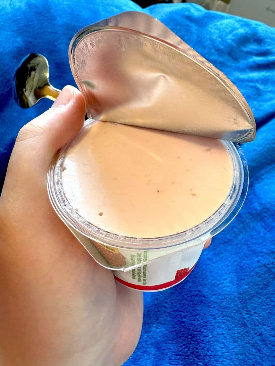 Hand holding a container of creamy yogurt with a foil lid partially peeled back.