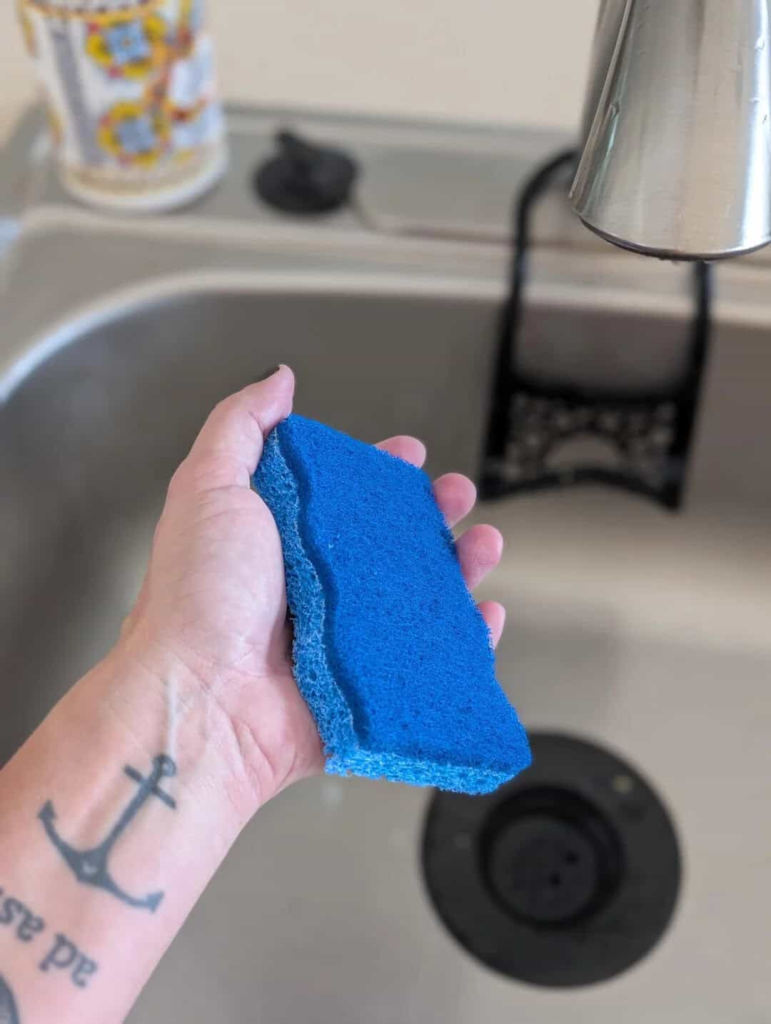 Hand holding a blue sponge near a kitchen sink with a faucet and dish soap bottle.