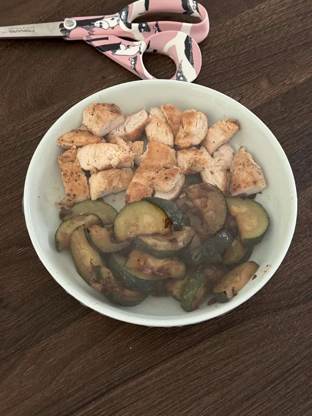 Chicken and zucchini in a white bowl on a dark wooden table with pink scissors nearby.