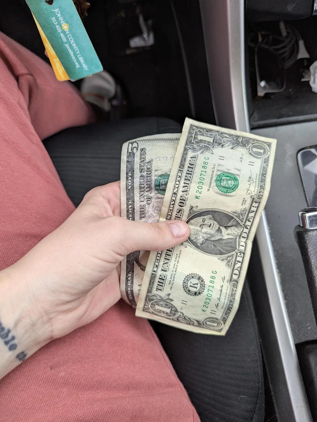 $20 and $1 bills held in a person's hand inside a vehicle.