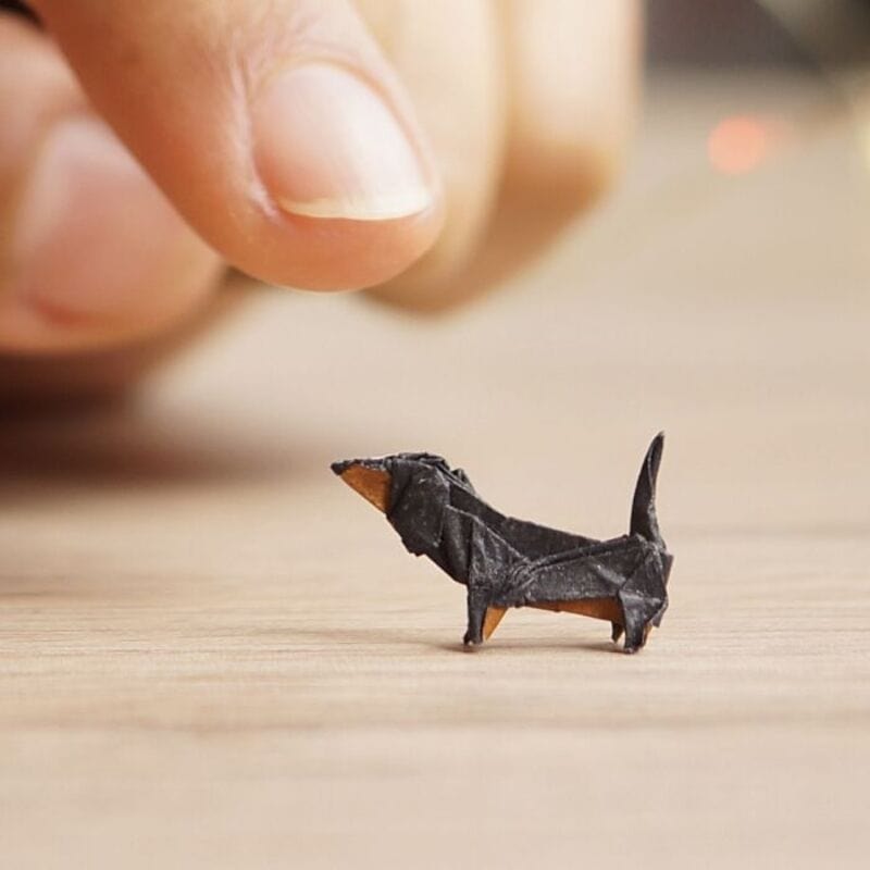 Miniature black paper dog with orange nose and tail, being held by a finger.