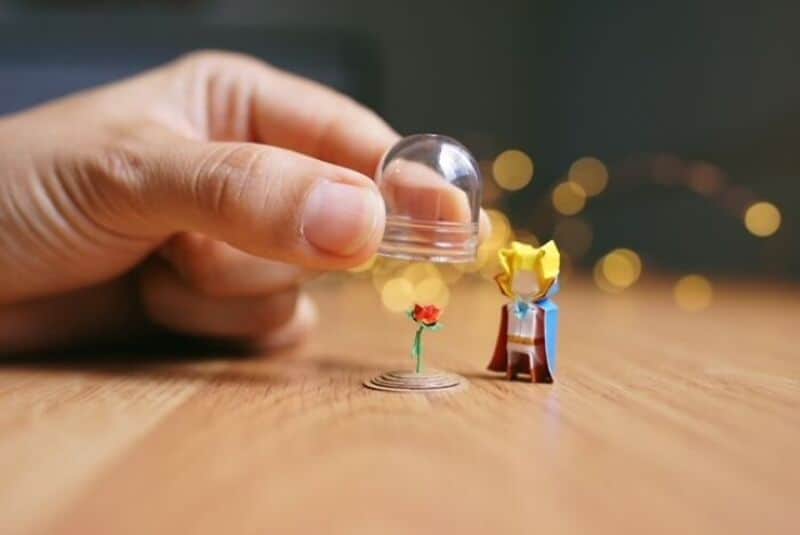 Hand placing glass dome over miniature paper art scene with a flower and a figure.