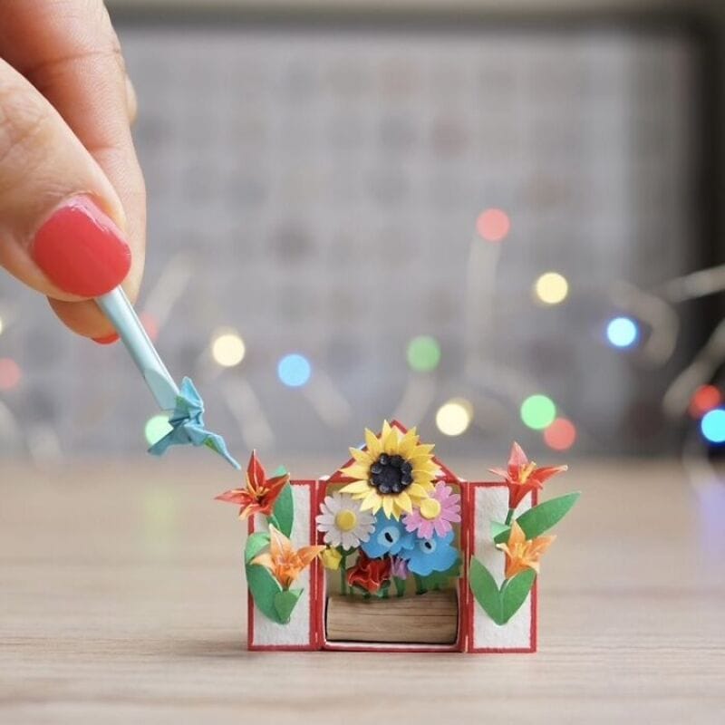 Miniature paper art of a flower garden with a hand holding a blue tool.