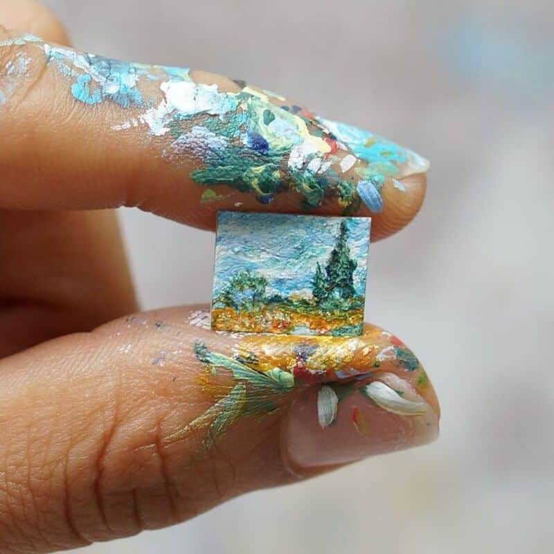 Miniature paper art of a landscape held between fingers with paint on fingers.