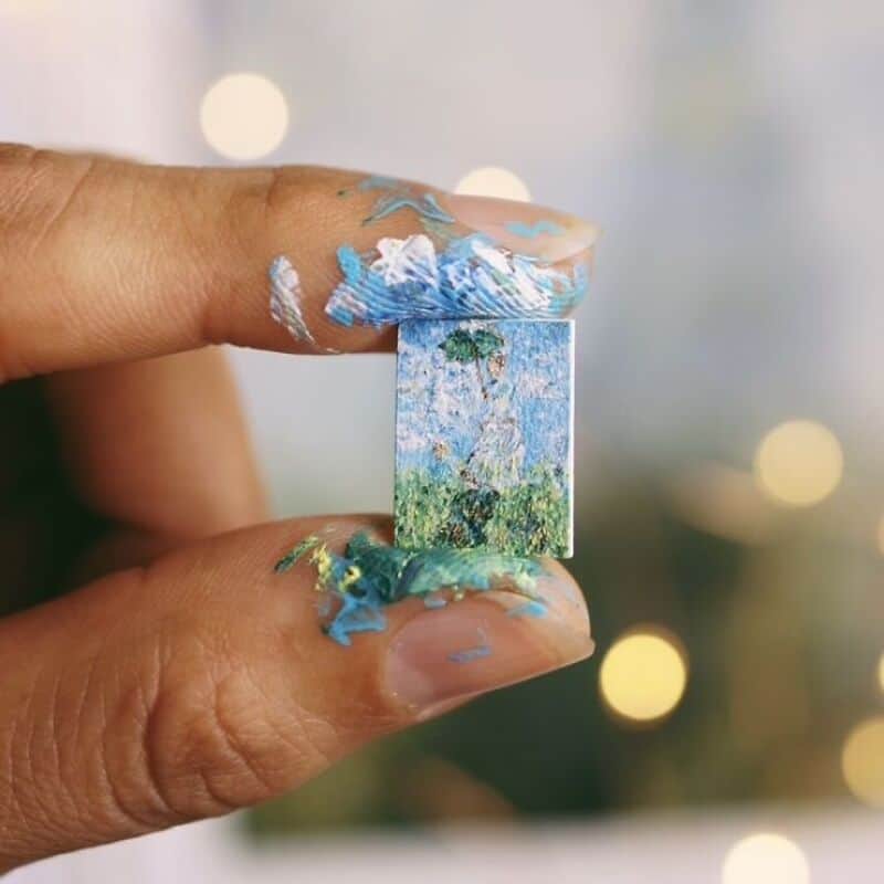 Miniature landscape painting held between fingers with paint smudges, blurred lights background.