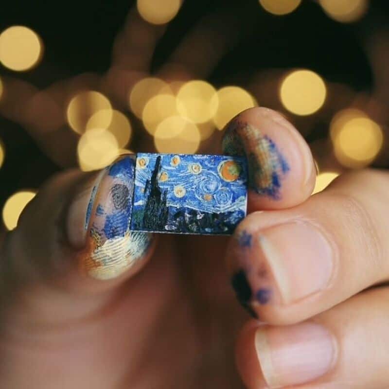 Miniature paper art piece held between fingers with painted nails, background of blurred lights.