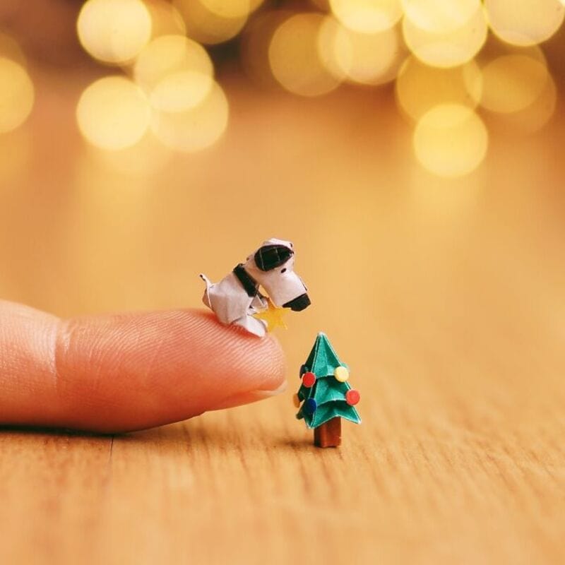 Miniature paper cow and Christmas tree on a wooden surface with blurred lights in background.