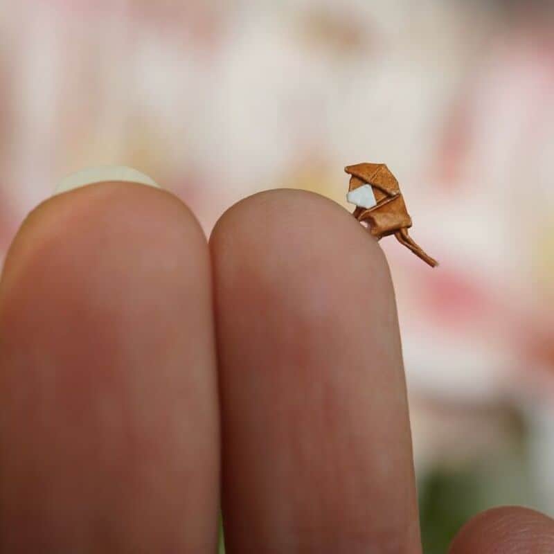 Miniature paper art of a tiny brown dog on a finger, with a blurred background.