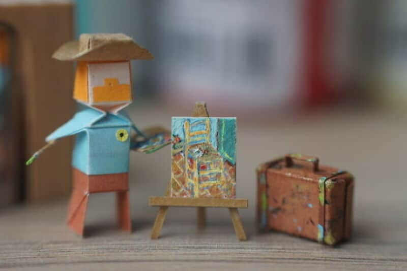Miniature paper art scene with a figure, easel, and suitcase on a wooden surface.