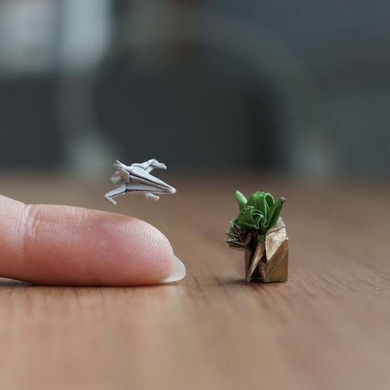 Miniature origami frog jumping towards a tiny paper plant on a wooden surface.