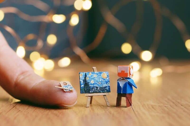 Miniature paper art scene with tiny easel, canvas, and figure, with blurred lights in background.