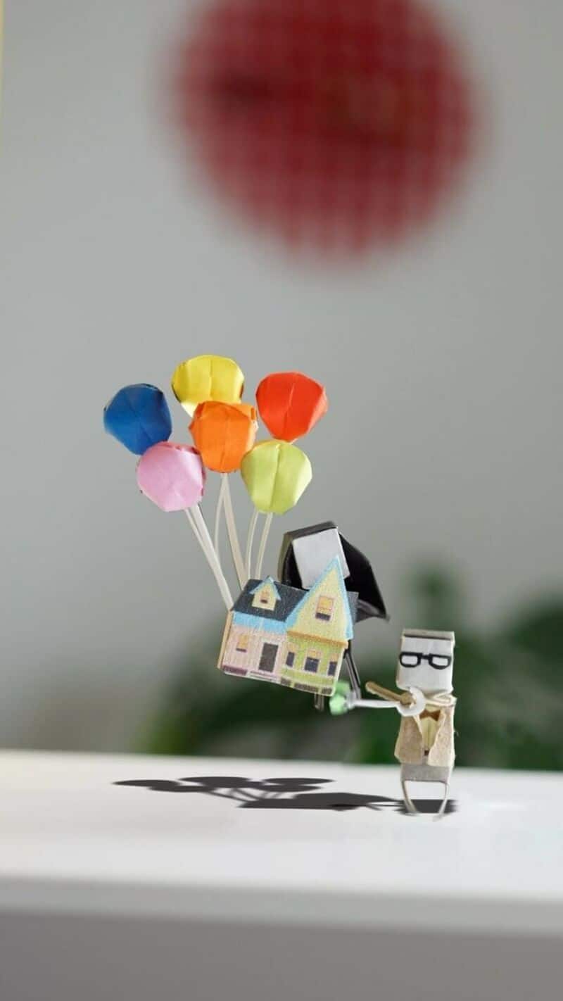 Miniature paper house with colorful paper balloons and a paper figure holding glasses.
