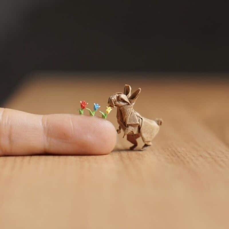 Origami rabbit with tiny colorful paper flowers on finger, wooden surface, blurred background.