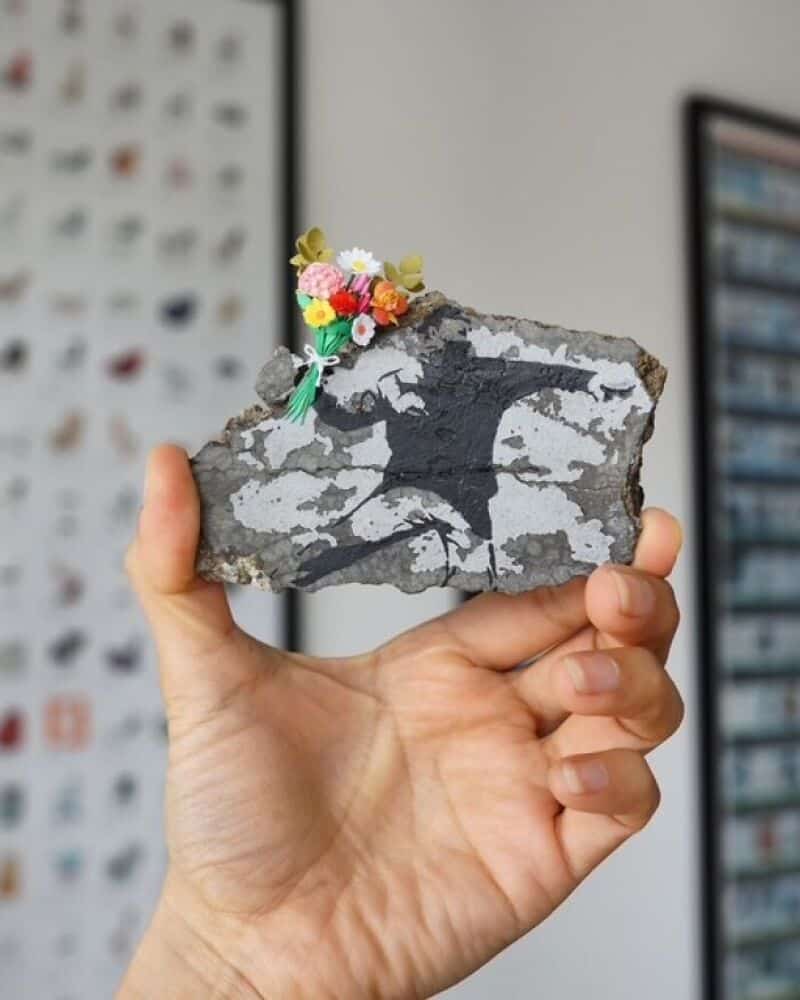 Miniature paper art of a black cat with flowers on its head, held in a hand.