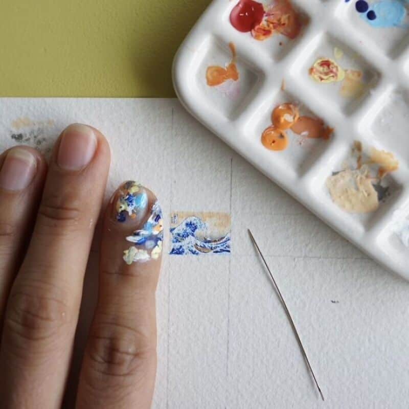 Close-up of a finger with blue and white nail art, watercolor palette, and fine brush.