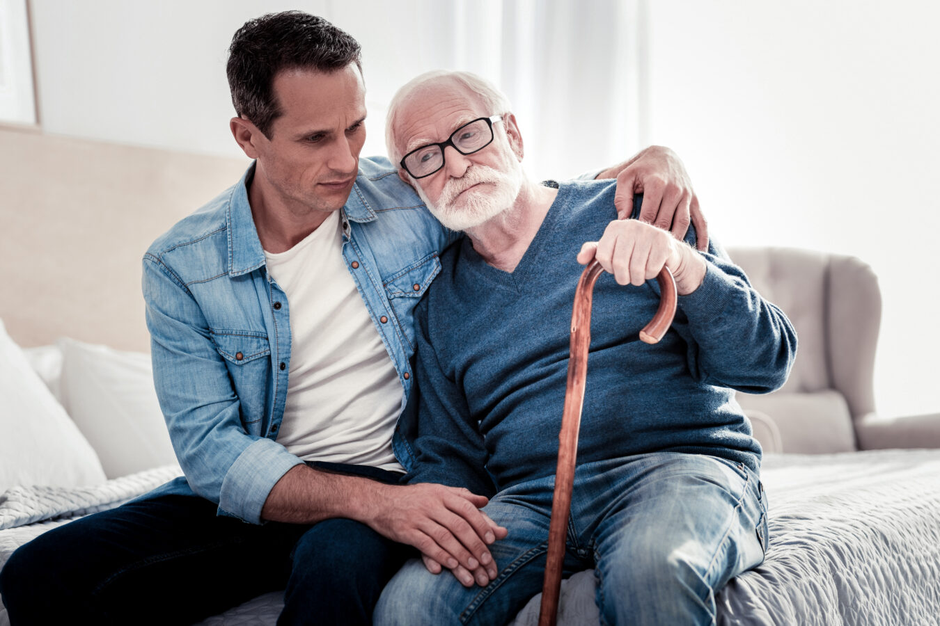 Elderly man with cane and caregiver sitting on bed, comforting each other.