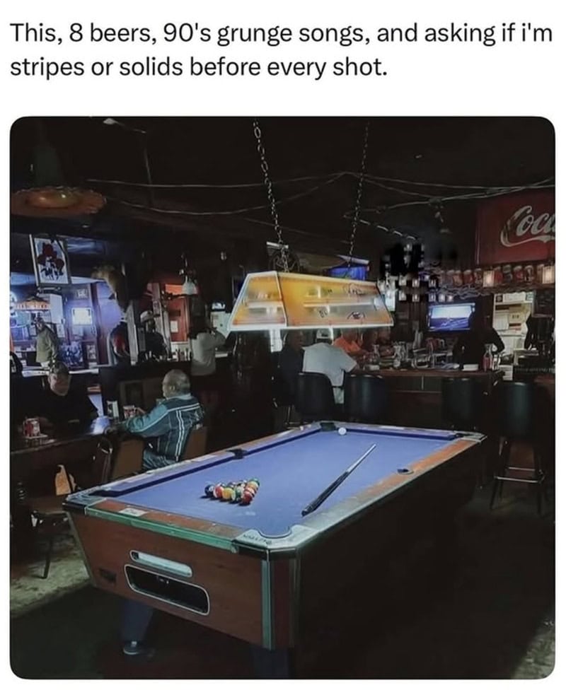 Pool table with balls and cue stick in a bar setting with people and arcade machines.
