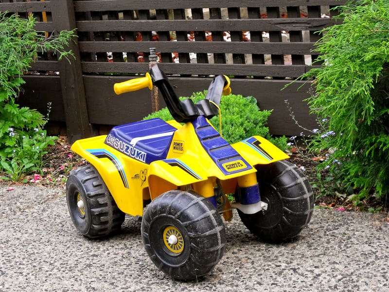 Yellow and blue children's ride-on toy quad bike on gravel with greenery.