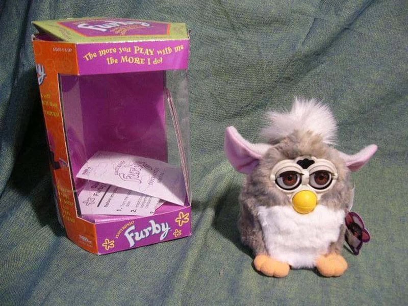 Furby toy with glasses and yellow nose next to its box on a green fabric surface.