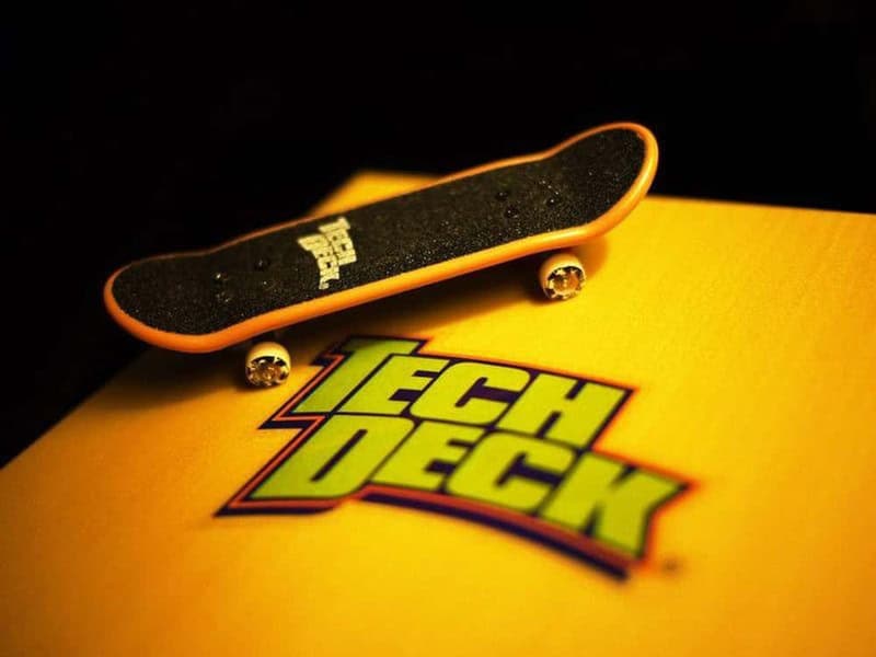 Mini skateboard on a yellow Tech Deck box with a black grip tape and orange edges.