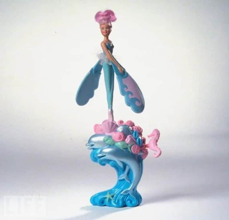 Toy figurine of a fairy with pink hair, blue dress, and large blue wings, standing on a colorful sea.