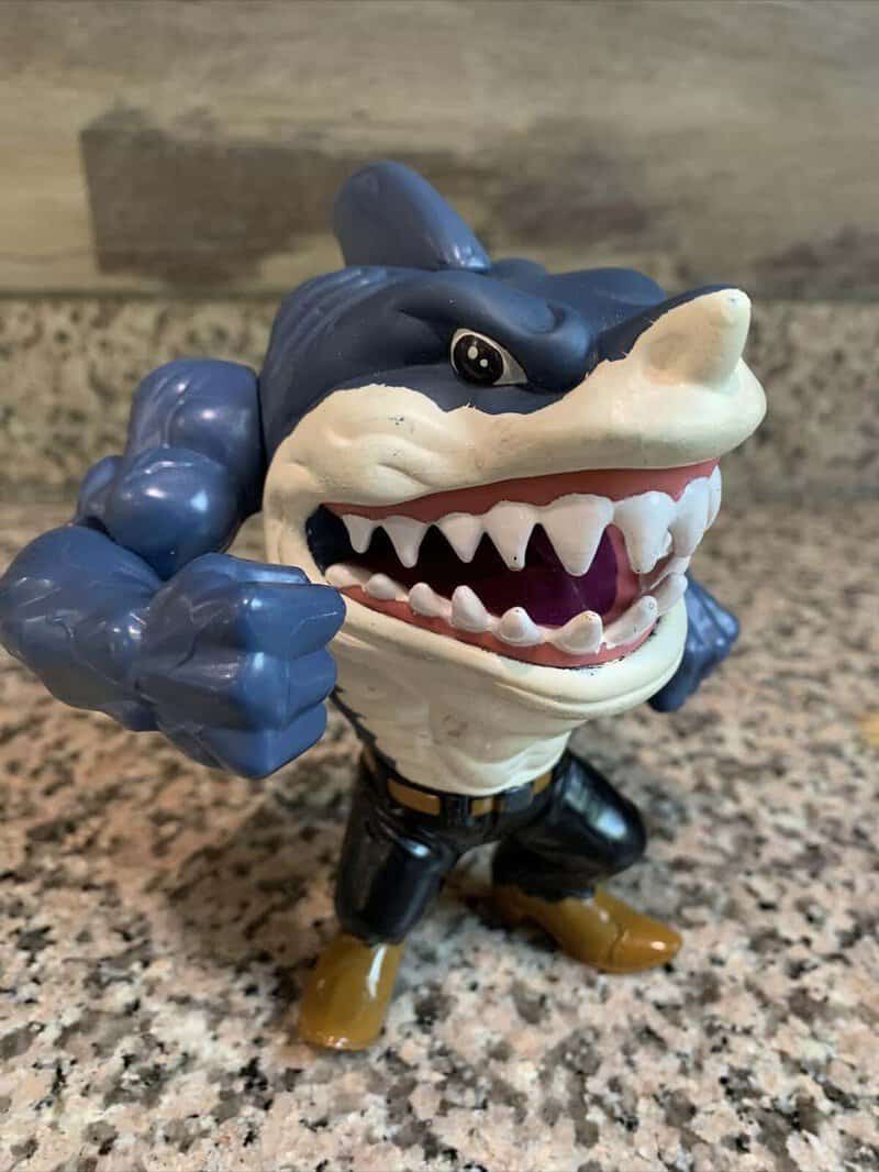 Toy figure of a blue shark with a humanoid body, wearing black pants and brown shoes.