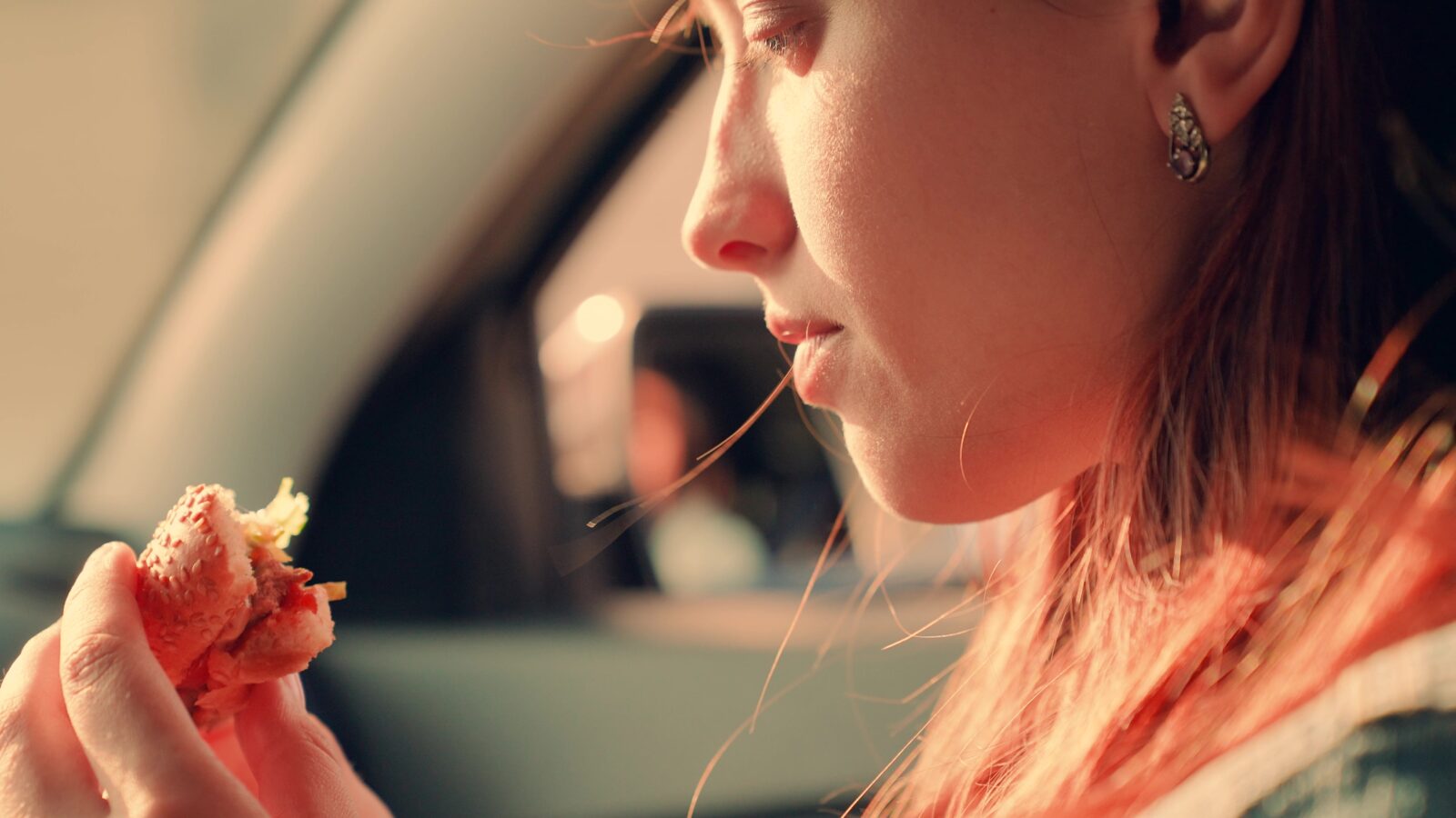 A woman with red hair eating a sandwich in a car.
