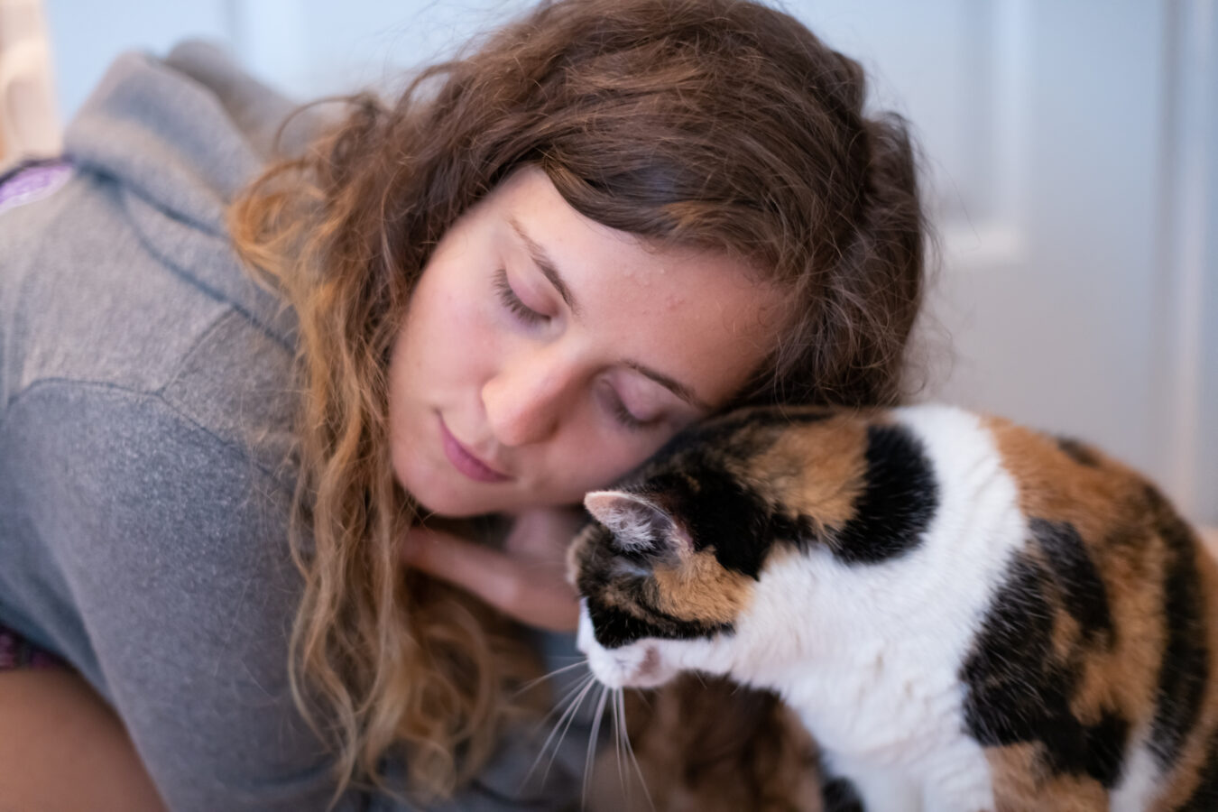 A woman with curly brown hair gently touching her face to a calico cat.