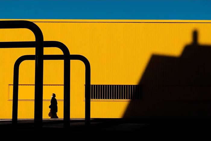 A woman walking past a yellow building with black pipes and shadows.