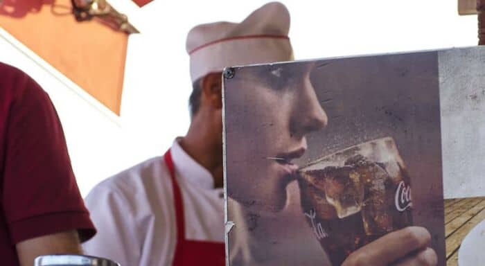 Coca-Cola drinker holding a glass of soda with a straw, partially obscured by a sign.