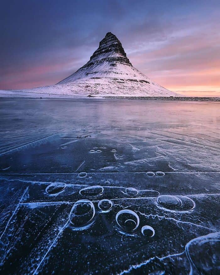 Snow-covered mountain with icy lake and bubbles in the foreground at sunset.