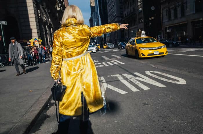 A woman in a shiny yellow coat crossing a busy city street with a yellow taxi approaching.