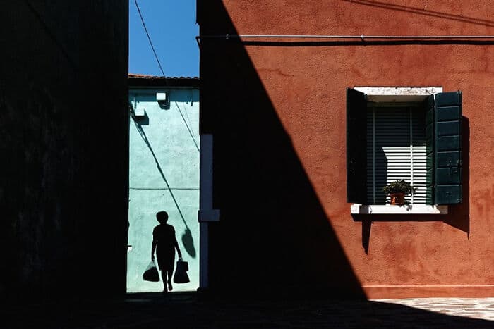 Silhouette of a person carrying bags walking past a colorful building with a window.