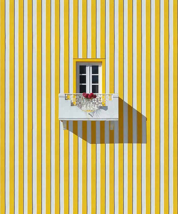 White balcony with flowers on a yellow and white striped wall, window above.