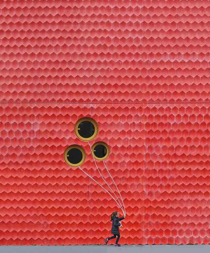 A girl running while holding three balloons against a red textured wall.