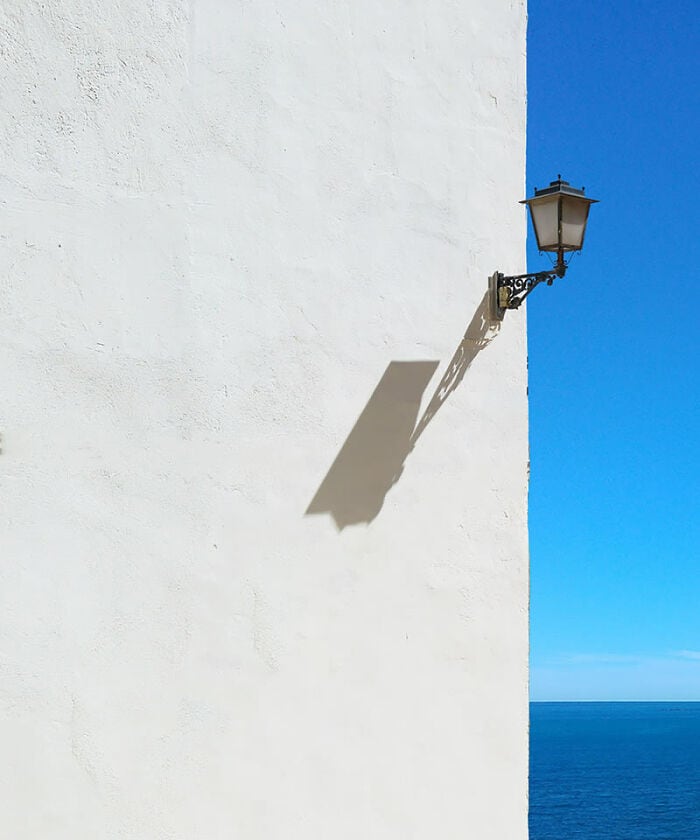 Vintage street lamp casting shadow on white wall by the sea under blue sky.