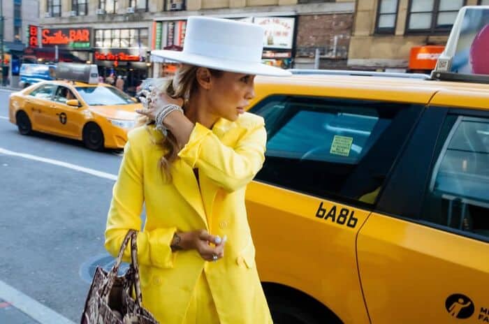 Yellow-clad woman with white hat on city street near yellow taxi cab.