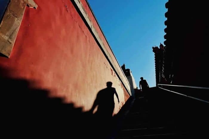 Shadows of two people on a red wall with stairs and a blue sky background.