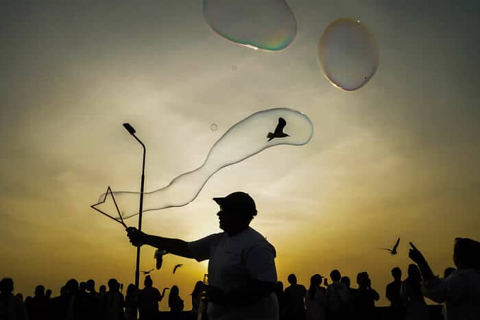 A man blowing bubbles at sunset with a crowd in the background.