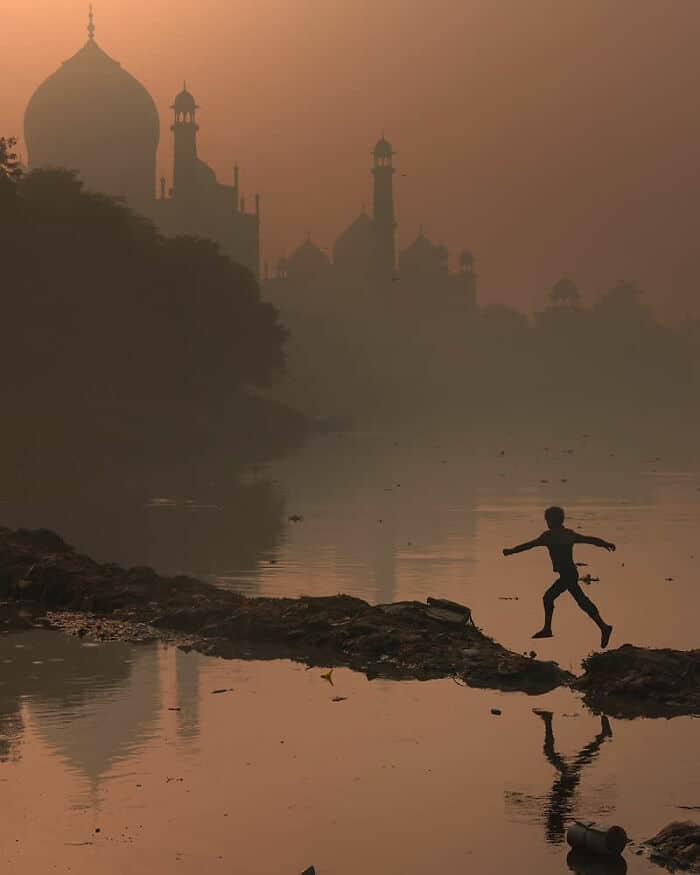 Child playing on a rocky riverbank with Taj Mahal in the background at sunset.