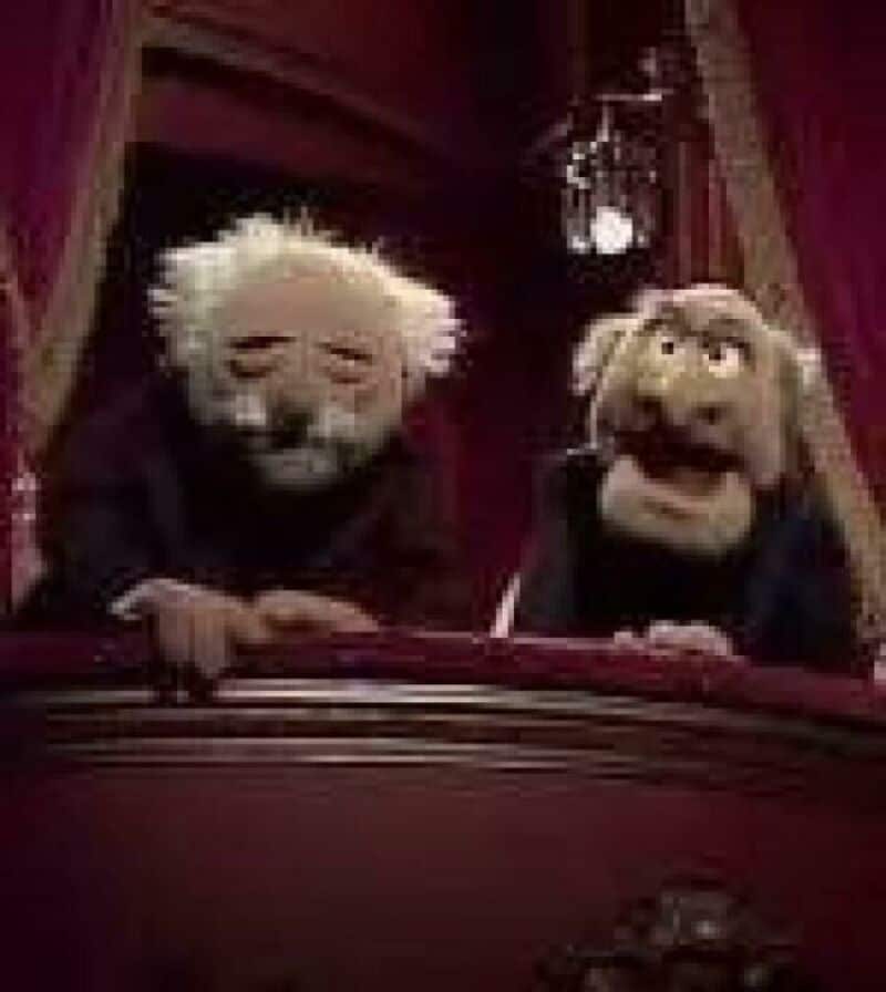 Two puppets with white and brown hair leaning over a theater balcony.